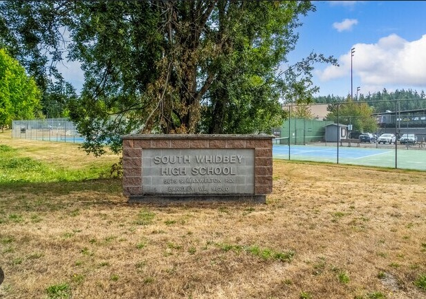 A brick-framed stone sign reading South Whidbey High School, Langley, WA, on a dry lawn, with tennis courts and a tall tree behind it.